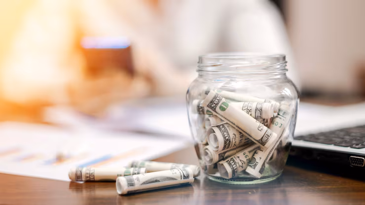 jar-with-rolled-banknotes-table-laptop-papers-woman-background_1268-17480